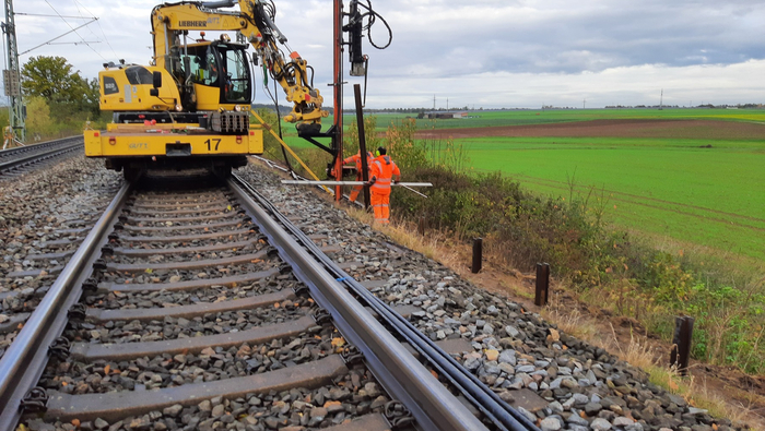 Bahnstrecke auf der ein Zweiwegebagger fährt. Rechts daneben zwei Menschen in orangener Schutzkleidung.