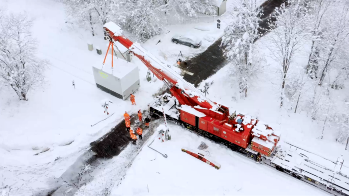 In einer stark verschneiten Landschaft ist ein Kran auf Bahngleisen und stellt einen weißen viereckigen Kasten neben die Strecke.